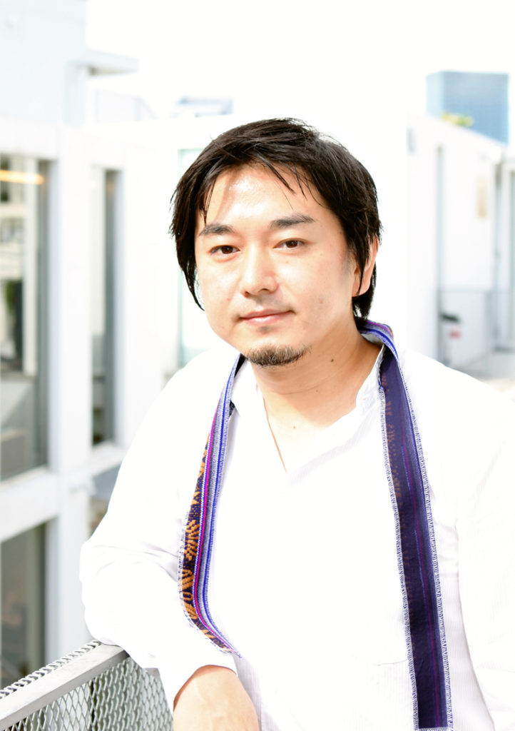 A colour photograph of Japanese architect Akihisa Hirata. He is wearing a white shirt open at the collar with a thin purple coloured scarf. He is leaning with his right arm on a grey metal balcony, and there are white window frames in the background.