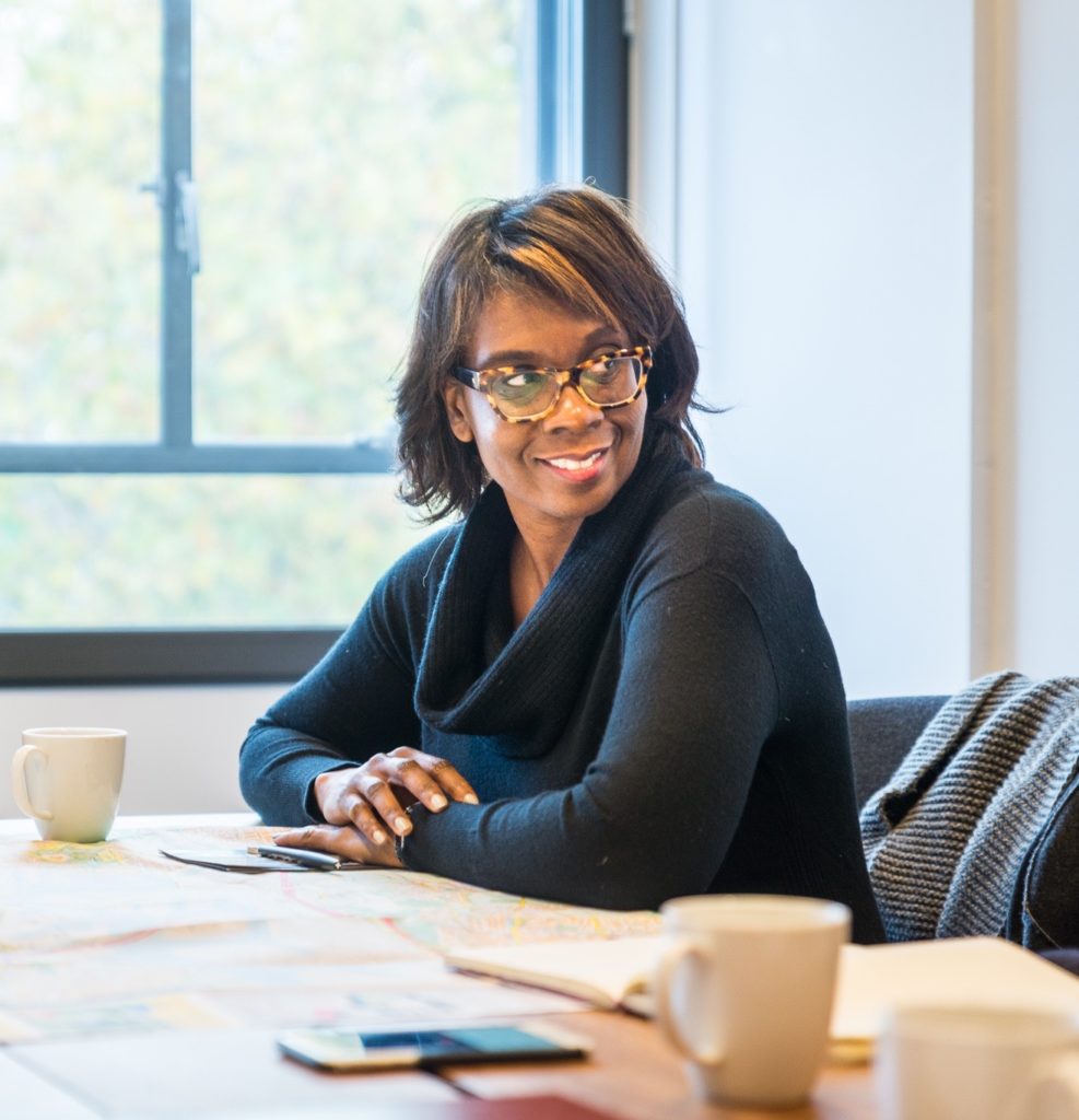 A colour photograph of Toni L. Griffin. She is seated at a table, with her arms slightly folder on the edge, and she is turning slightly to look over her shoulder, smiling. She is wearing glasses and a dark blue sweater. There is a white cup on the table in front of her alongside various papers. In the background there is a window with greenery outside.