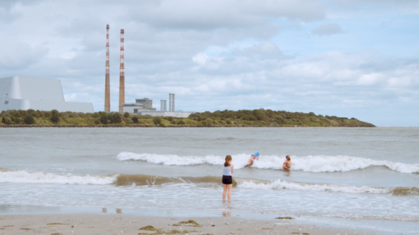 A still from the Site Specfic short film about Poolbeg Towers. The image is in colour and shoes the beach at Sandymount Strand with the red and white striped chimneys visible to the left of the picture. There are some figures splashing and paddling in the sea at the centre of the image. The sky is mostly blue with some clouds.