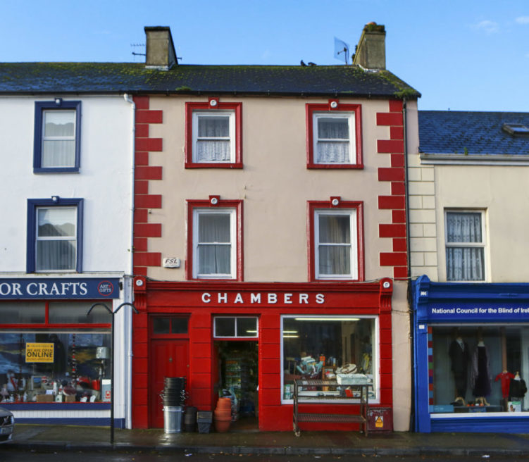 A photograph taken of a street in Kilrush. The photo shows three shopfronts, brightly painted in primary colours. Chambers' shop with a red shopfront is at the centre. The sky is blue.
