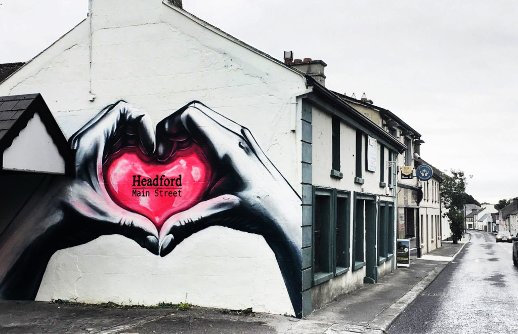 Photo of a mural painted on the end of a row of buildings in Headford. The mural shows hands forming a heart shape around a pink heart with the words 