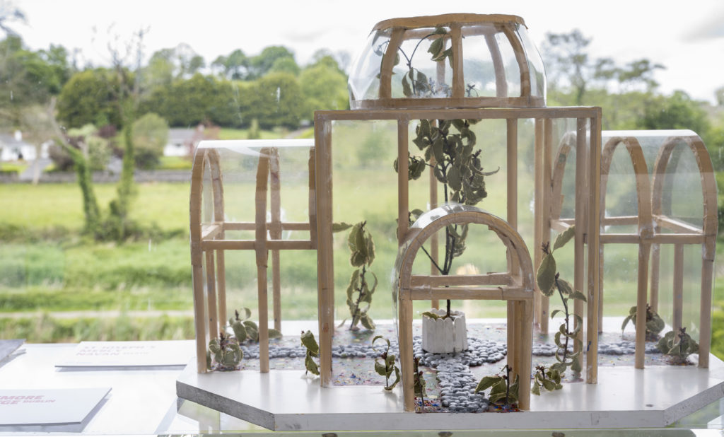 Close-up photo of a 3D model of an outdoors classroom, which resembles a nineteenth-century glasshouse. It is on display on a window ledge, and the verdant Museum grounds can be seen through it and the glass, so it appears to be in the landscape.