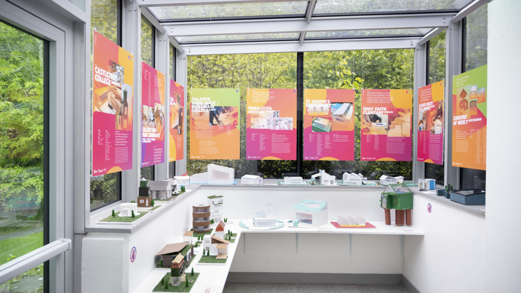 Photo of AIS Exhibition 3D models on 2 shelves, with brightly coloured posters hanging in front of the glass walls above.