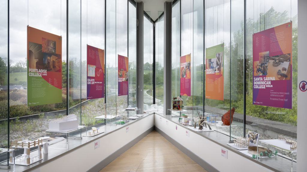 Photo of AIS Exhibition posters and 3D models displayed on 2 glass walls that meet at an acute angle, overlooking the grounds of the Museum.