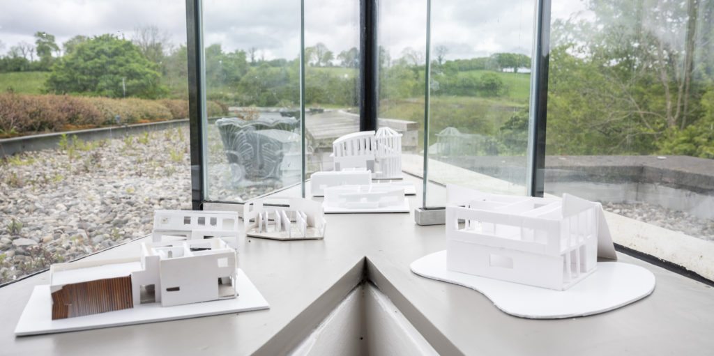 Close-up photo of four white 3D models on the low window ledge in the acute corner where two glass walls meet. The ground and trees are visible through the glass behind them, so that the models almost appear as buildings in the landscape.