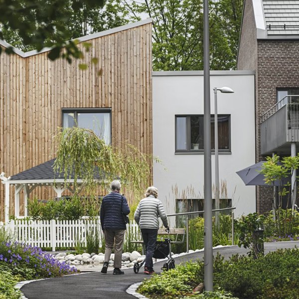 Photo by Benjamin A. Ward of the Carpe Diem Dementia Village in Oslo. An elderly man and woman with their backs to the camera walk down a path towards modern 2-storey buildings. There is a gazebo and gardens in front of the buildings.