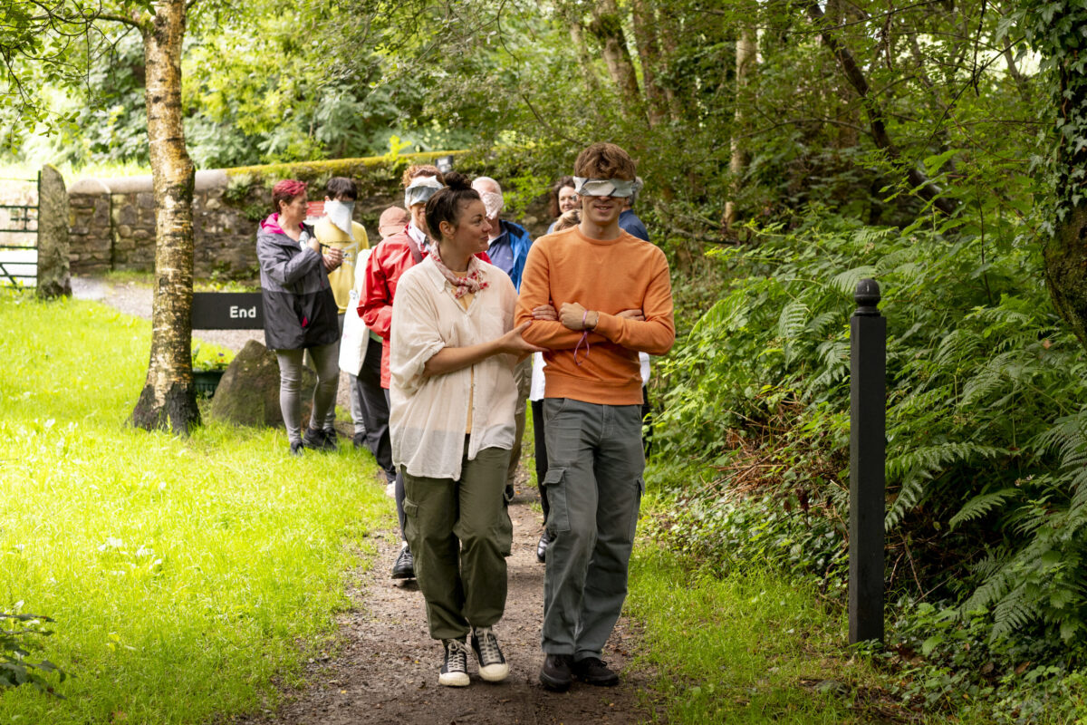 On a sunny day, a group of people are walking in twos down a path between lush grass, trees, and bushes. One person in each pair is blindfolded and the other is leading them. There is a stone wall and gate in the background.